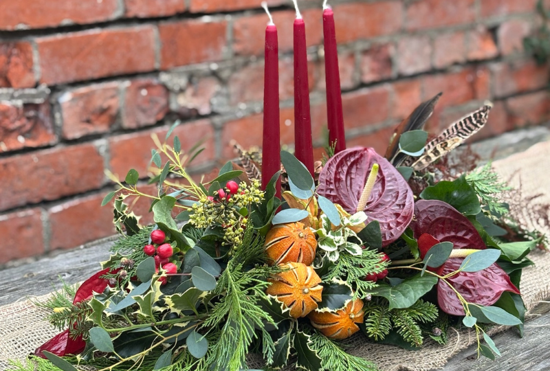 Noël Luxe Christmas table arrangement with red candles anthuriums dried oranges berries pine foliage and a pheasant feather displayed on a rustic table.